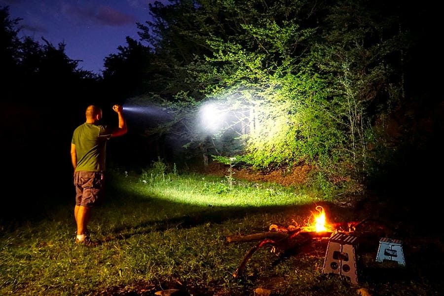 Man shining flashlight into woods beside a campfire