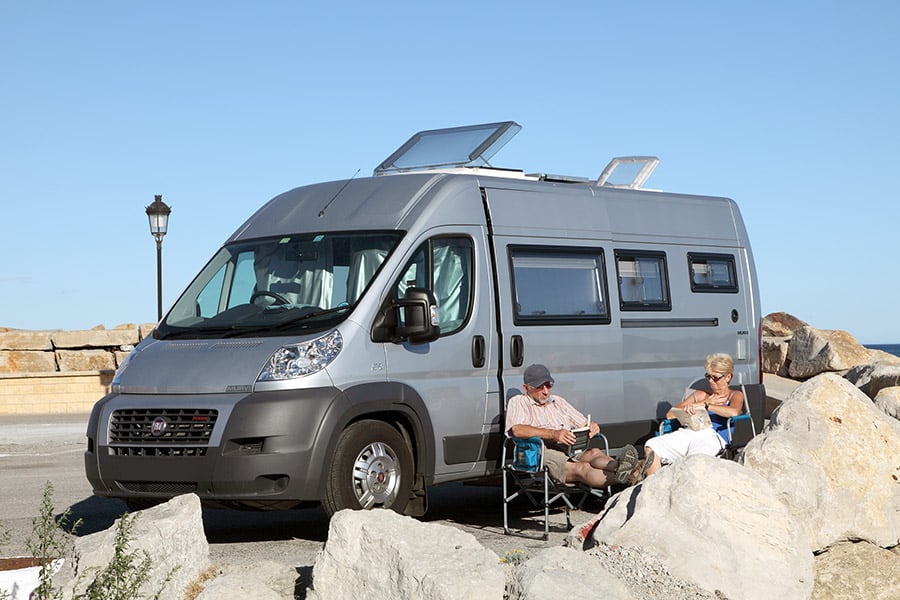 A couple sitting in camping chairs beside a gray camper van with big rocks in foreground