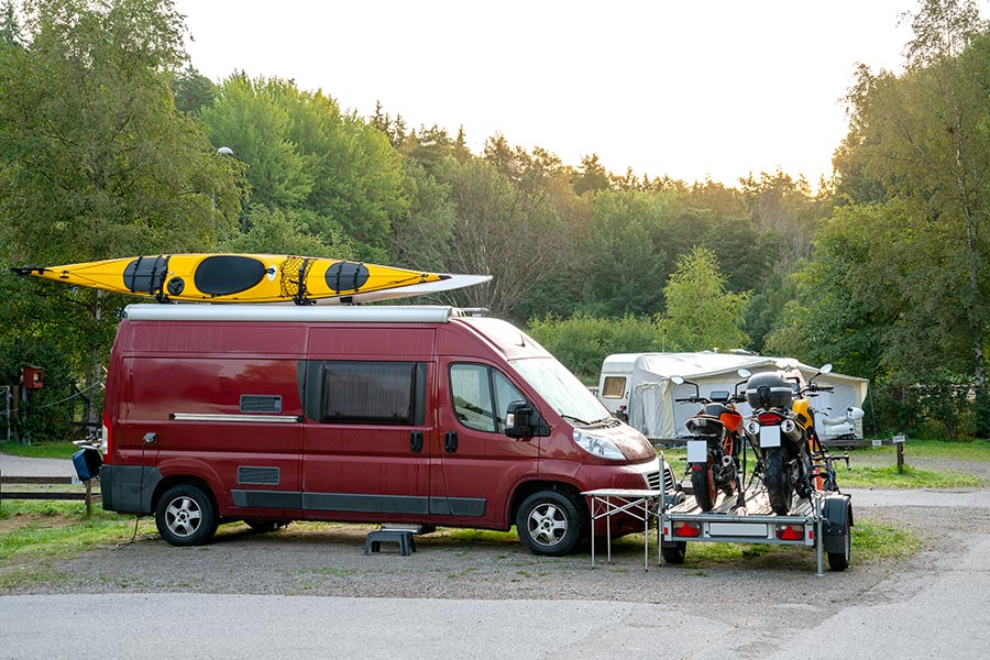 Red camper van with a yellow kayak on top and motorcycles parked on a trailer in front of it