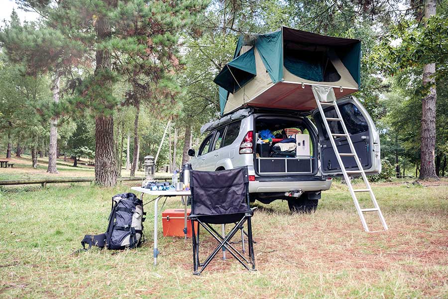 Silver SUV with a roof tent parked beside a table and chairs in a wooded area