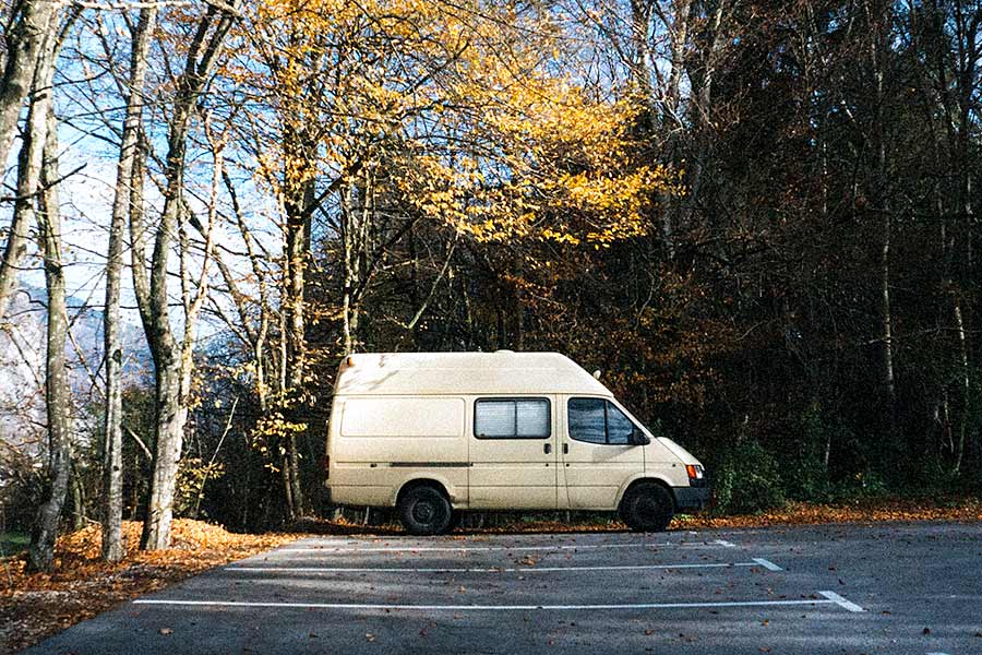 Light colored camper van parked in a parking lot with trees behind it