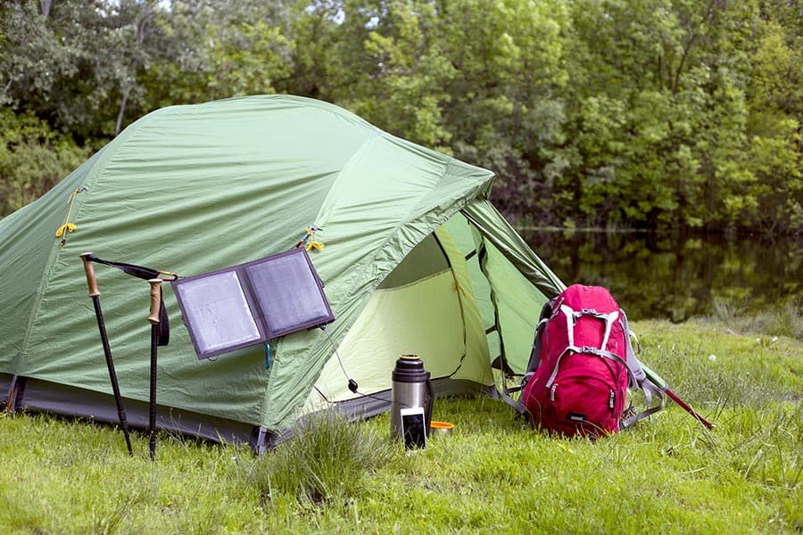 Green tent in a wooded area with backpack, hiking poles and a portable solar panel in front of it
