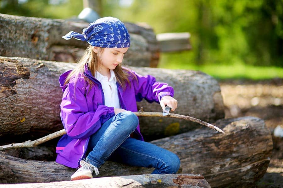 Girl in purple coat sitting on log whittling a stick