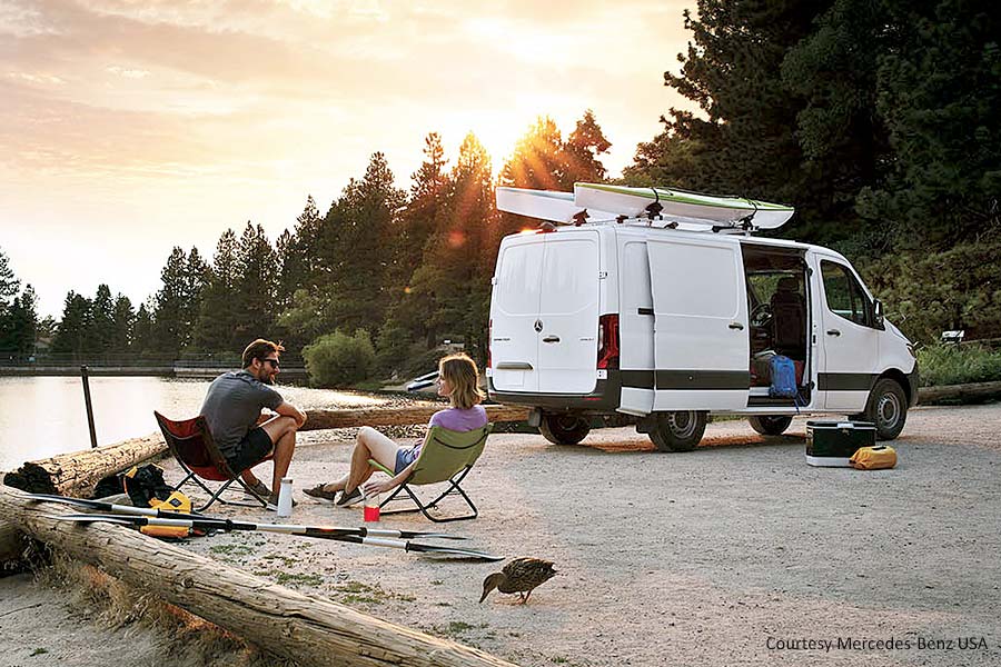 A white van parked by the water with two people in camping chairs behind it