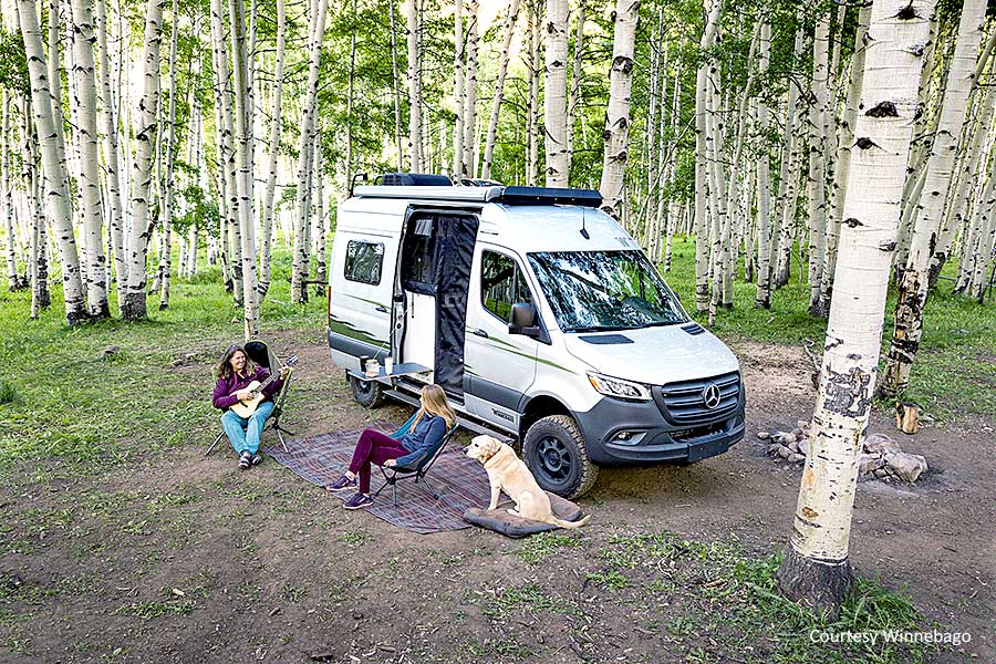 White camper van parked in woods with campers in chairs relaxing beside it