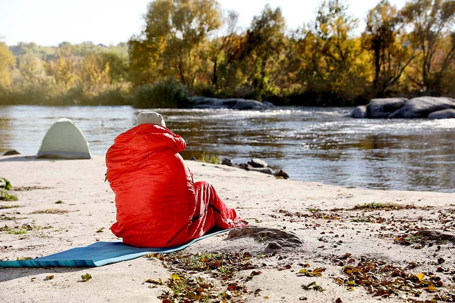 Man in sleeping bag sitting on mat by rivers edge