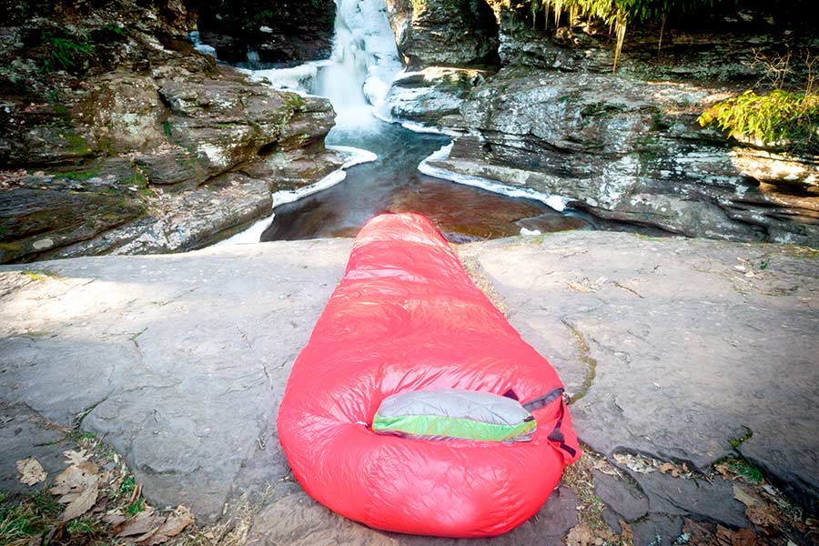 Red mummy sleeping bag laying on rock ledge by water