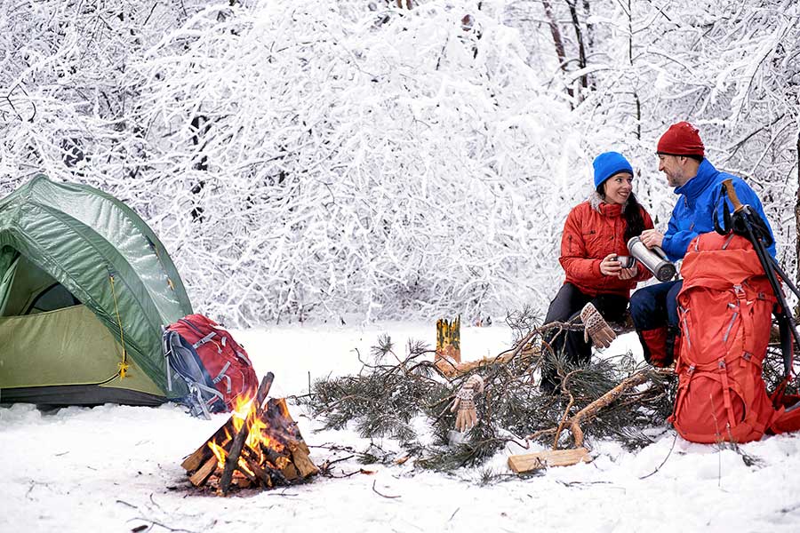 Couple winter camping, sitting by fire drinking hot coffee