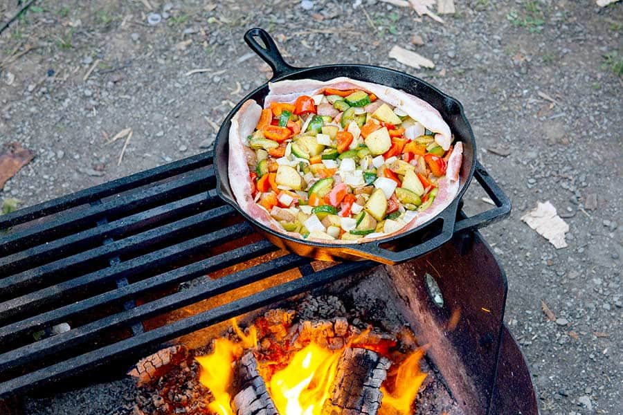 Cast iron skillet full of vegetables and meat cooking over campfire